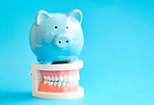 Light blue piggy bank sitting on top of a set of dentures representing the cost of dental implants